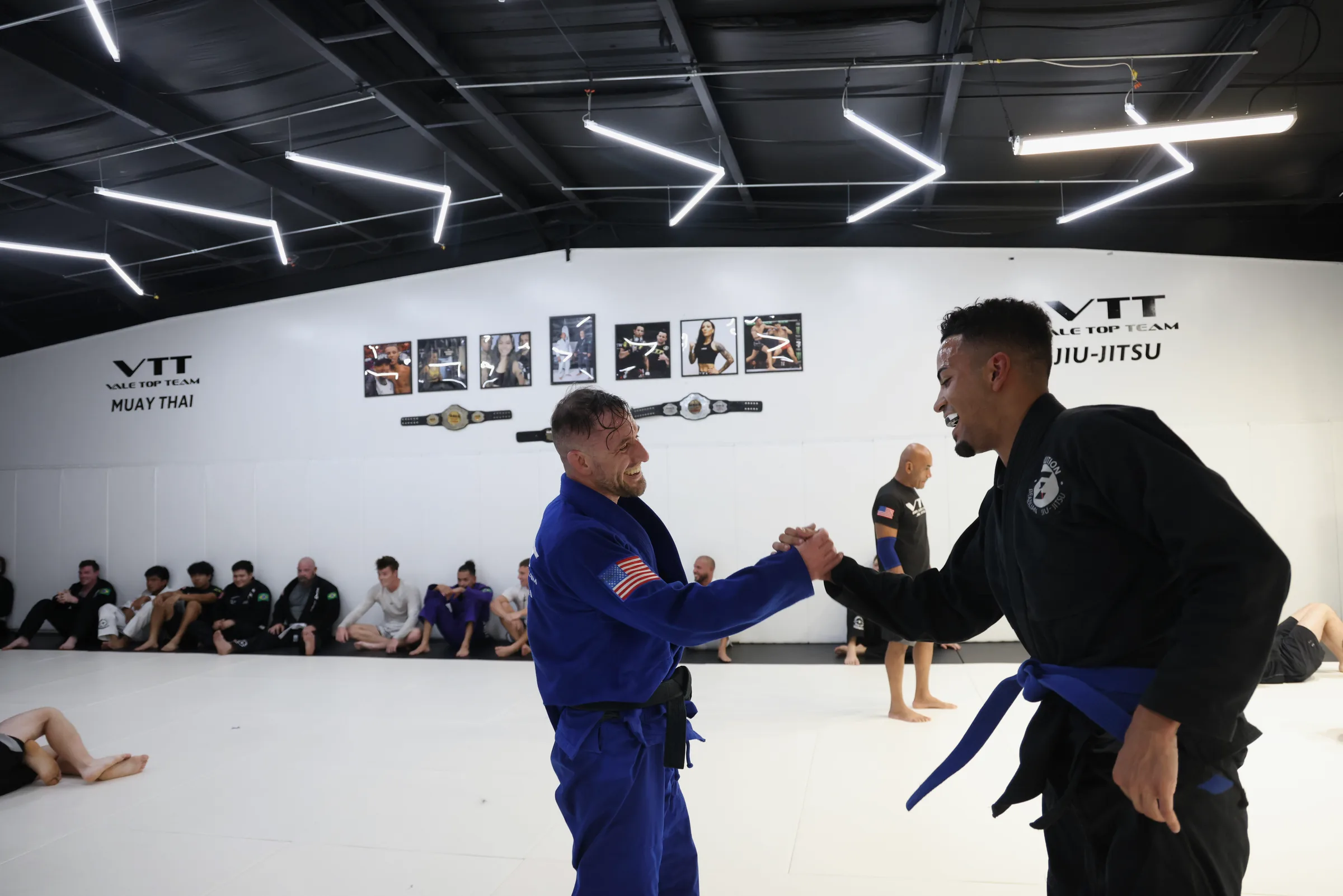 A wide-angle view of a jiu-jitsu academy training floor. Cj, in a blue gi with black belt, stands mid-handshake with a training partner in a black gi with blue belt. Both are smiling. Other practitioners are seated along the wall in the background, watching. Bright institutional lighting reflects on the white mat and walls.