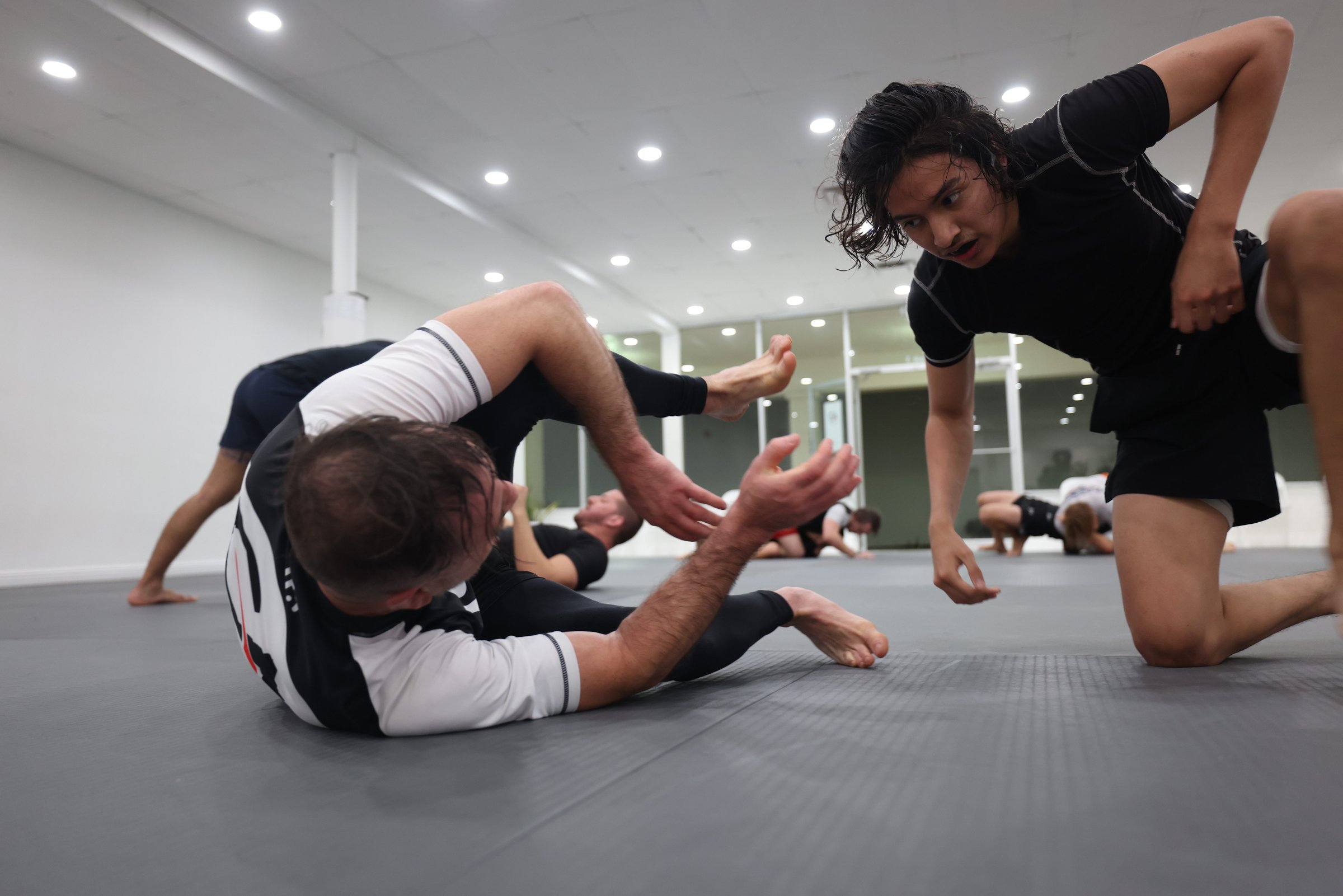 Three jiu-jitsu practitioners training on a grey mat under institutional lighting. One in the foreground recovers on his back, hand reaching upward. Another leans in to engage. The image is honest, unromanticized, mid-roll.